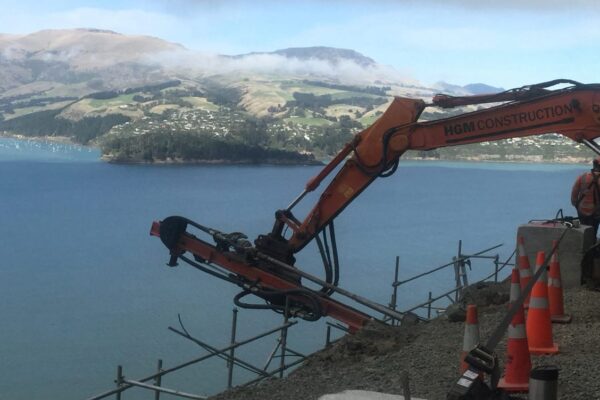 Panoramic image of an excavator-mounted construction drill working beside a harbour road during civil construction and anchoring works. This image shows civil contractors delivering drilling services, rock anchors, soil nails, slope retention and ground stabilisation in a coastal environment.