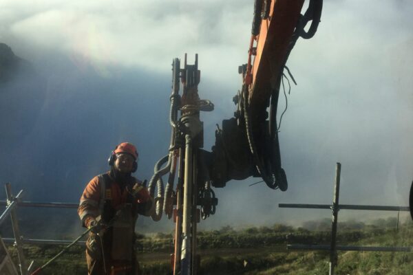 Civil contractor monitoring a construction drill during rock anchor installation on a steep coastal site above the harbour. The image shows drilling services, anchoring and piling, slope retention, soil nails and ground stabilisation as part of a complex civil construction project.