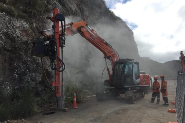 Excavator-mounted construction drill working against a rocky roadside slope, with civil contractors supervising drilling services for slope retention and ground stabilisation. This image highlights rock anchors, soil nails, retaining walls preparation and civil construction on a constrained coastal road.