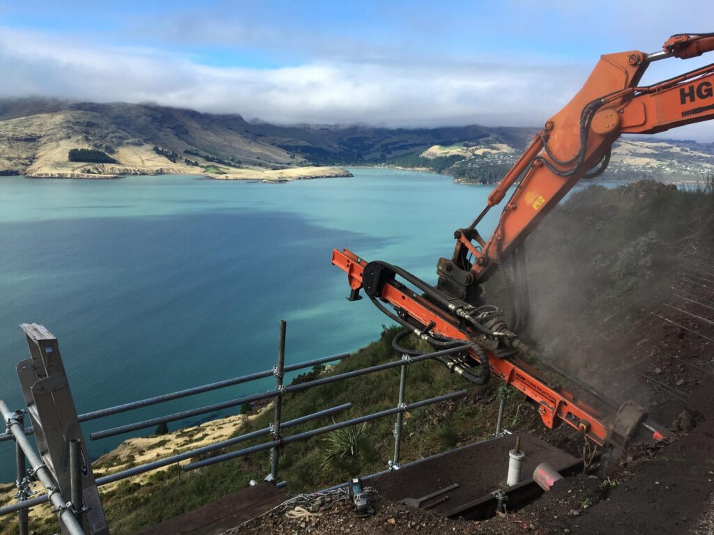Civil construction and drilling services underway on a steep coastal hillside, with an excavator-mounted construction drill installing rock anchors above the harbour. This image highlights slope retention, ground stabilisation, site construction and complex construction projects managed by civil contractors.