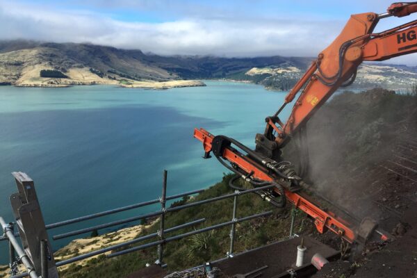 Civil construction and drilling services underway on a steep coastal road, with an excavator-mounted construction drill completing rock anchors for slope retention above the harbour. The image shows civil contractors managing ground stabilisation, site construction and complex construction projects in a constrained hillside environment.