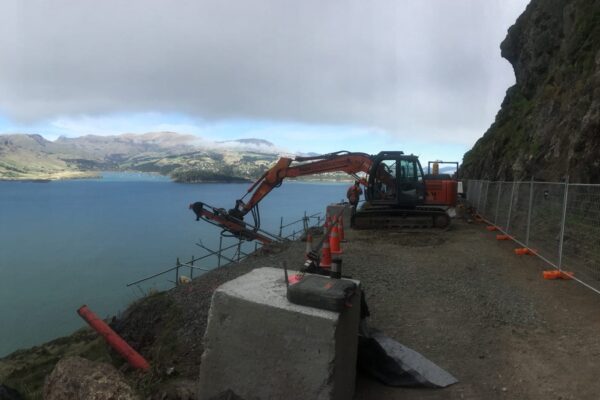 Excavator and construction drill set up on a narrow hillside road for civil construction, slope retention and ground stabilisation works. The image shows civil contractors carrying out drilling services, rock anchors and siteworks above the coastline.