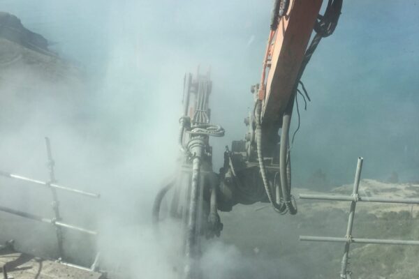 Construction drill operating on a steep coastal slope during civil construction and anchoring works, with excavation equipment supporting drilling services above the harbour. This image shows civil contractors completing rock anchors, soil nails, slope retention and ground stabilisation on a difficult access site.
