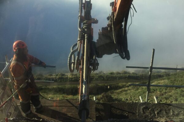 Civil contractor standing beside a construction drill during rock anchor installation on a steep coastal road above the harbour. The image highlights drilling services, anchoring and piling, slope retention, ground stabilisation and site construction for a complex construction project.