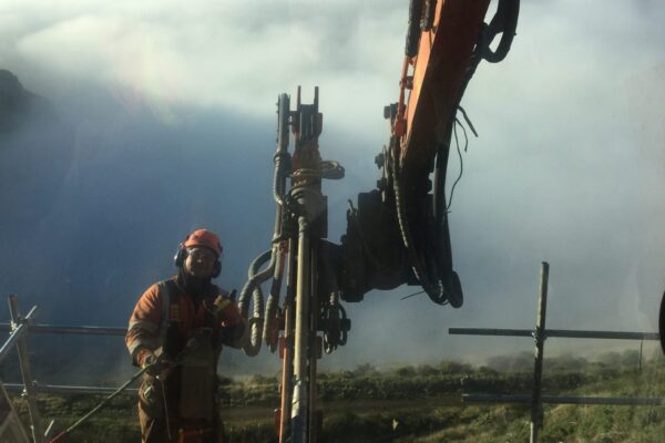 Drilling services and rock anchor installation taking place on a hillside construction site, with civil contractors working beside an excavator-mounted construction drill. This image shows slope retention, ground stabilisation, anchoring and piling, and civil construction above a coastal landscape.