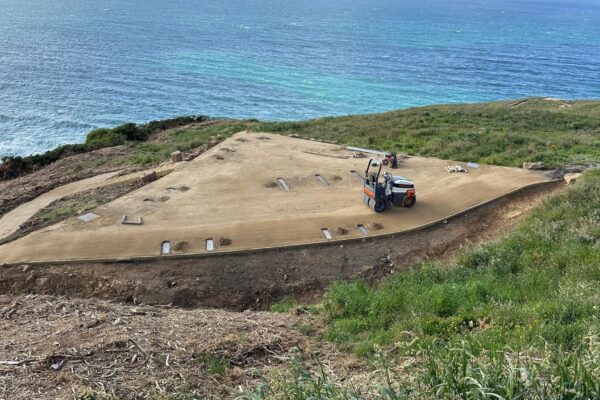 Coastal walking track construction with a compaction machine finishing a wide gravel pathway above the shoreline. The project shows civil construction, recreation construction, site preparation and erosion control across a steep coastal site.