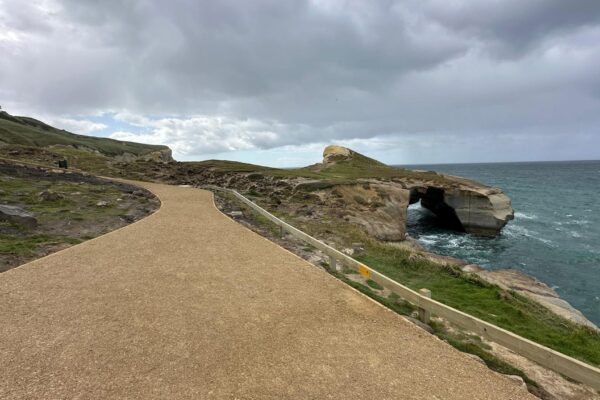 Completed walking track construction along a rugged coastal landscape, with a stabilised gravel path overlooking sea cliffs and natural rock formations. This civil construction project includes recreation construction, erosion control and coastal construction work for public access.