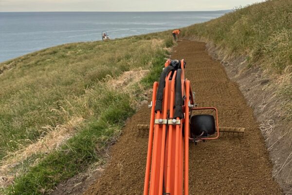 Excavation machinery working along a narrow coastal path during walking track construction above the ocean. This recreation construction project includes civil construction, siteworks, erosion control and careful land development on a steep coastal slope.