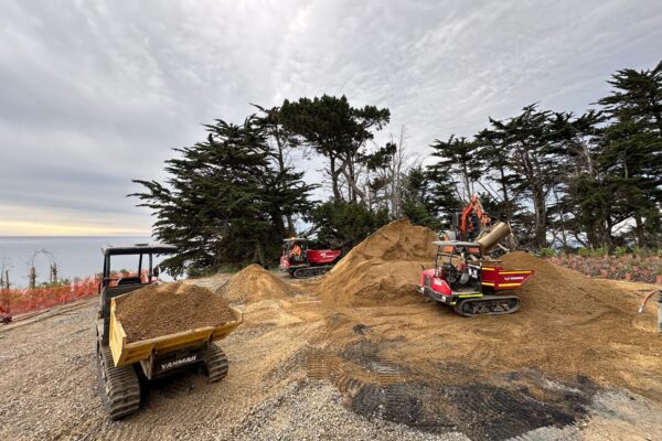 Earthworks contractors and compact machinery shaping gravel paths and stockpiles for walking track construction near the coast. The image shows civil construction, site preparation, erosion control and recreation construction in a coastal environment.