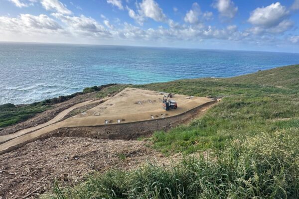 Coastal walking track construction with a small compaction machine working on a newly formed gravel path above the shoreline. This civil construction project includes recreation construction, siteworks, erosion control and land development work on exposed coastal terrain.
