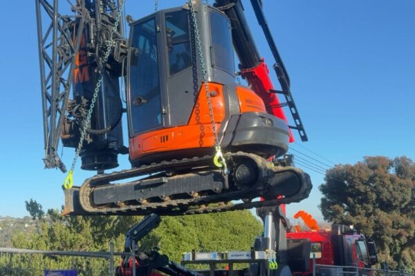 Excavator being lifted by crane truck during sling operations for access to a restricted civil construction site. The image shows machinery transport, site preparation and project management for retaining walls and excavation works.