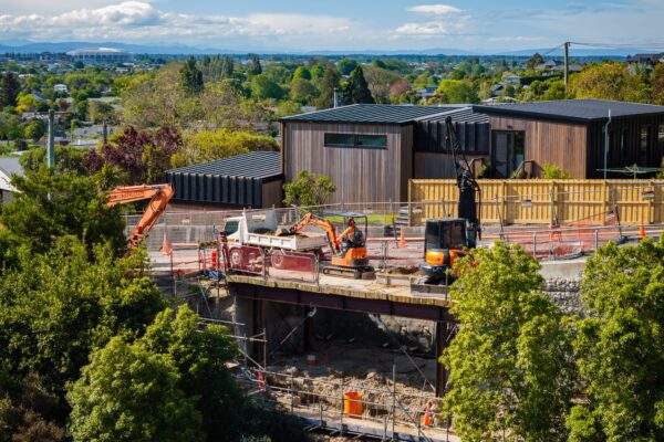 Civil construction machinery working beside a residential hillside site with retaining walls, scaffolding and safety barriers in place. The image shows excavation, structure construction and project management on a complex construction project.