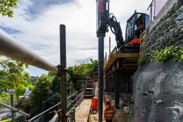 Civil contractors working below a construction drill on a steep excavation site for retaining walls and slope retention. The image shows specialist drilling services, rock anchors and concrete structure work forming part of a complex construction project.