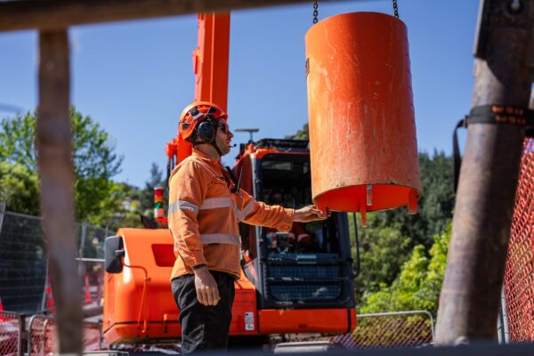 Civil construction worker overseeing sling operations as a concrete structure component is lifted beside excavation machinery. The image highlights retaining walls work, construction drill access and site contractors managing a complex construction project.