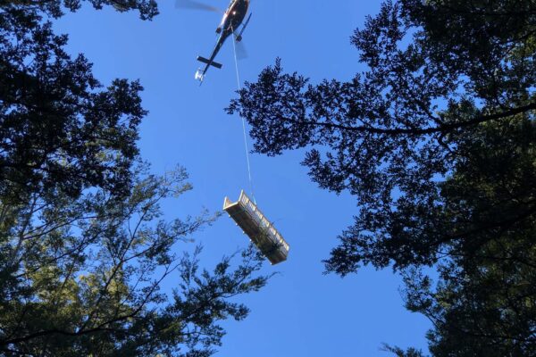 Helicopter operation lifting materials over a forested recreation construction site, supporting walking track construction and bridge construction in difficult access terrain. The image shows heli operation, commercial sling work and project management by civil contractors.