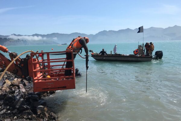 Marine construction crew completing harbour works from the shoreline and a small boat, with civil contractors managing marine excavation and coastal construction. This image shows marine services, utility infrastructure, siteworks and project management in a challenging marine environment.