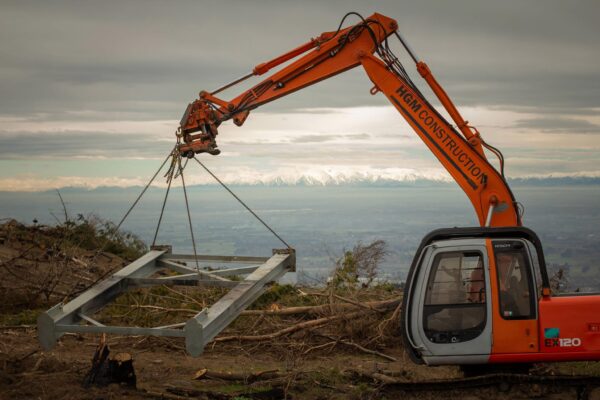 Excavator completing sling operations on a hillside recreation construction site, lifting steel components for civil construction and structure construction. The image shows civil contractors managing adventure infrastructure, machinery transport, project management and park construction in difficult access terrain.