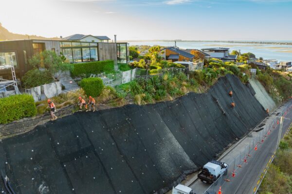 Aerial view of slope retention and rockfall mesh installation above a coastal road, with civil contractors working across a large stabilised bank. The image highlights retaining walls, civil construction, erosion control, ground stabilisation and large scale retaining walls.